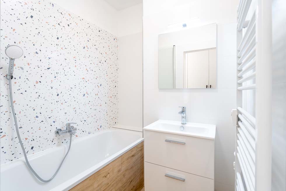 Salle de bain contemporaine dotée d'un élégant mur carrelé en terrazzo, d'un élégant lavabo blanc, d'une baignoire avec pomme de douche à main et d'un sèche-serviettes, créant un espace propre et accueillant